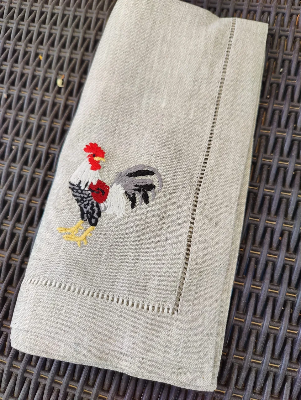 Servilleta de lino crudo doblada sobre ratán, con el gallo gris bordado de otro modelo