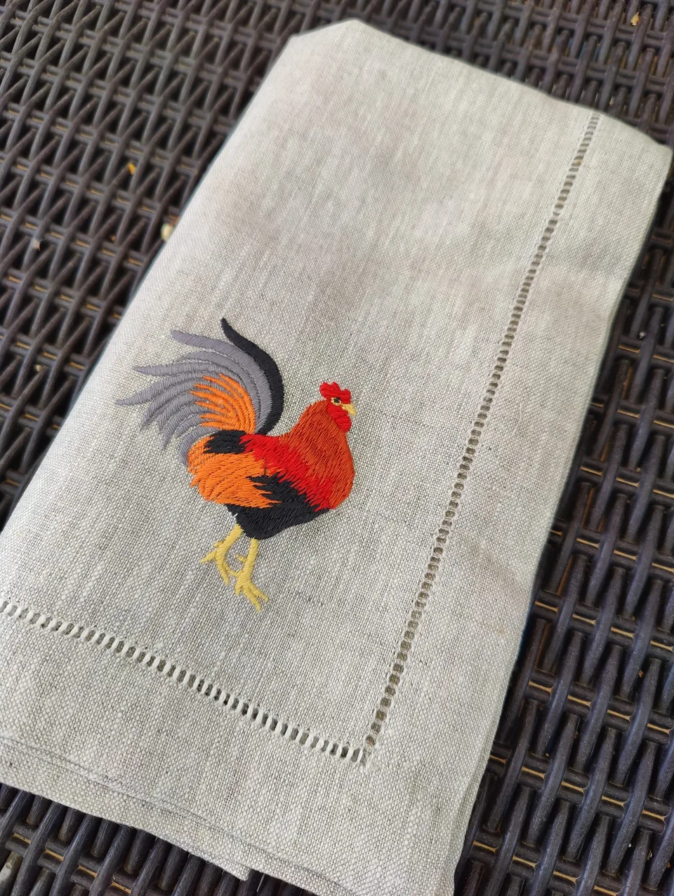 Servilleta de lino crudo doblada sobre ratán, con el gallo rojo bordado en la esquina