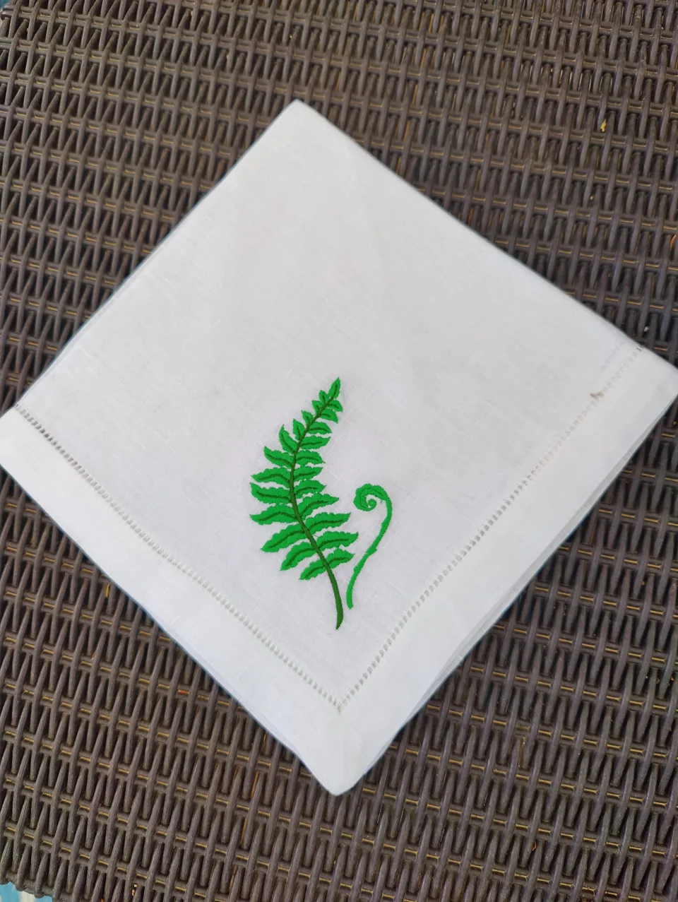 Servilleta de lino blanco con vainica y una hoja de helecho verde bordada, doblada sobre una silla de ratán