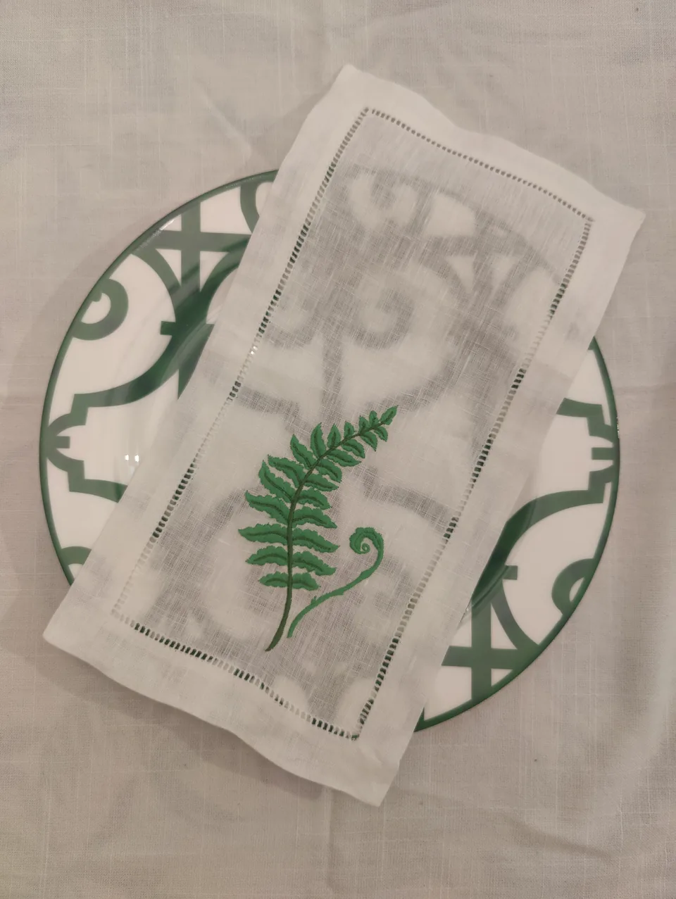 Servilleta de cóctel de lino blanco con vainica y una hoja de helecho verde bordada, sobre un plato de Rejas verde